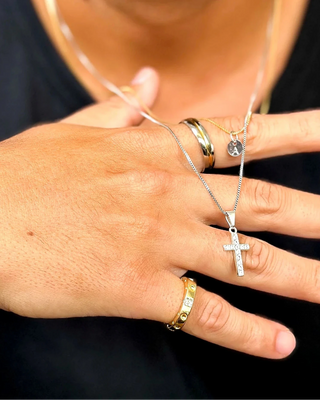 Aliança masculina de aço inox com filete IP gold da UNA Semijoias e Aço no dedo em close das mãos, com anéis e colares masculinos em destaque sobre camiseta preta.