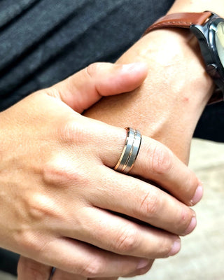Anel masculino de aço bicolor nas cores prata e rosé da UNA Semijoias e Aço no dedo, em close das mãos com relógio de couro marrom e look casual.
