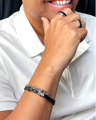 Anel masculino de aço inox 316L com faixa preta central da UNA Semijoias e Aço no dedo, em close do sorriso e do braço com pulseira masculina sobre camisa branca.