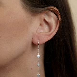 Argolinha com fio de coração banhada a prata 925 da UNA Semijoias e Aço na orelha feminina em close, com três corações pendentes em destaque.