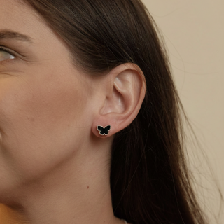 Brincos pequenos formato borboleta na cor preta, banhados a prata 925 da UNA Semijoias e Aço, em fundo branco minimalista com foco frontal nas peças.